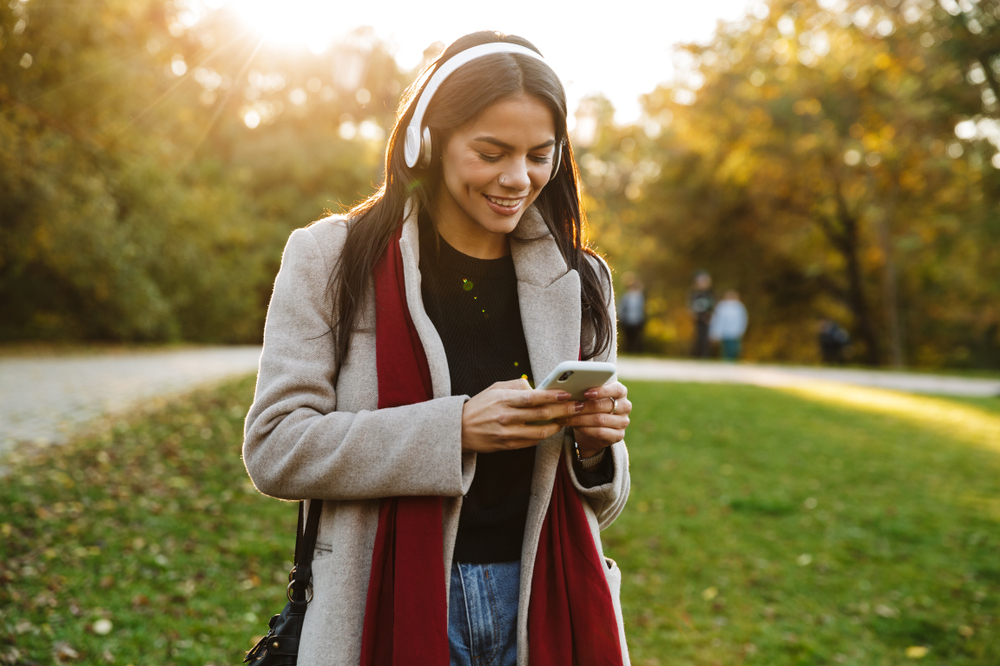 Portrait of happy nice woman wearing coat listening to music with headphones and using cellphone while walking in autumn park